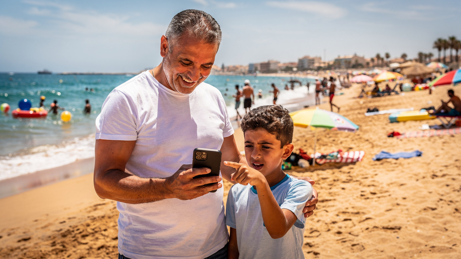 Un père montre l'écran de son téléphone à son fils, sur une plage animée d'Oran, le fils pointe du doigt l'écran avec un air curieux, symbolisant la découverte et la connectivité.
