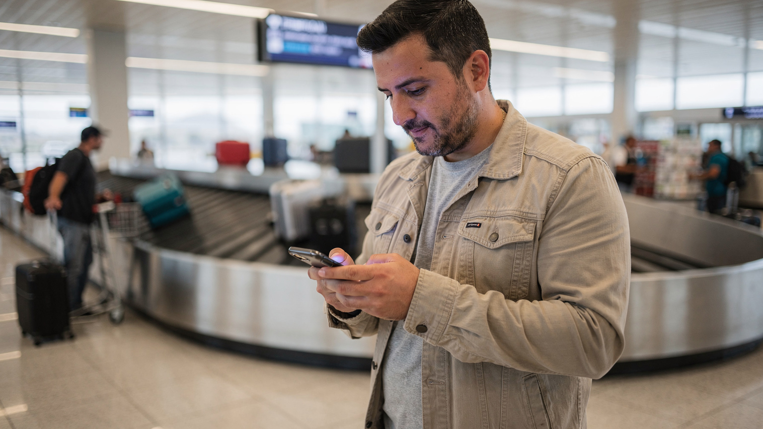 Un viajero consulta su teléfono, donde se ve la aplicación de Cellesim con un plan de eSIM activo, mientras espera su equipaje en la cinta del aeropuerto de Santiago.