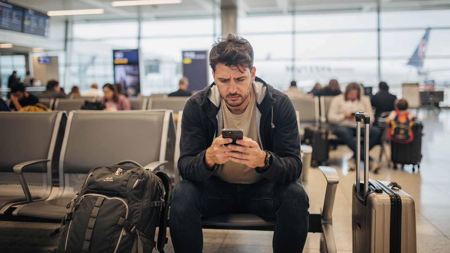 Un viajero cansado en una sala de espera de aeropuerto concurrida, mirando con preocupación su smartphone, con maletas a sus pies.