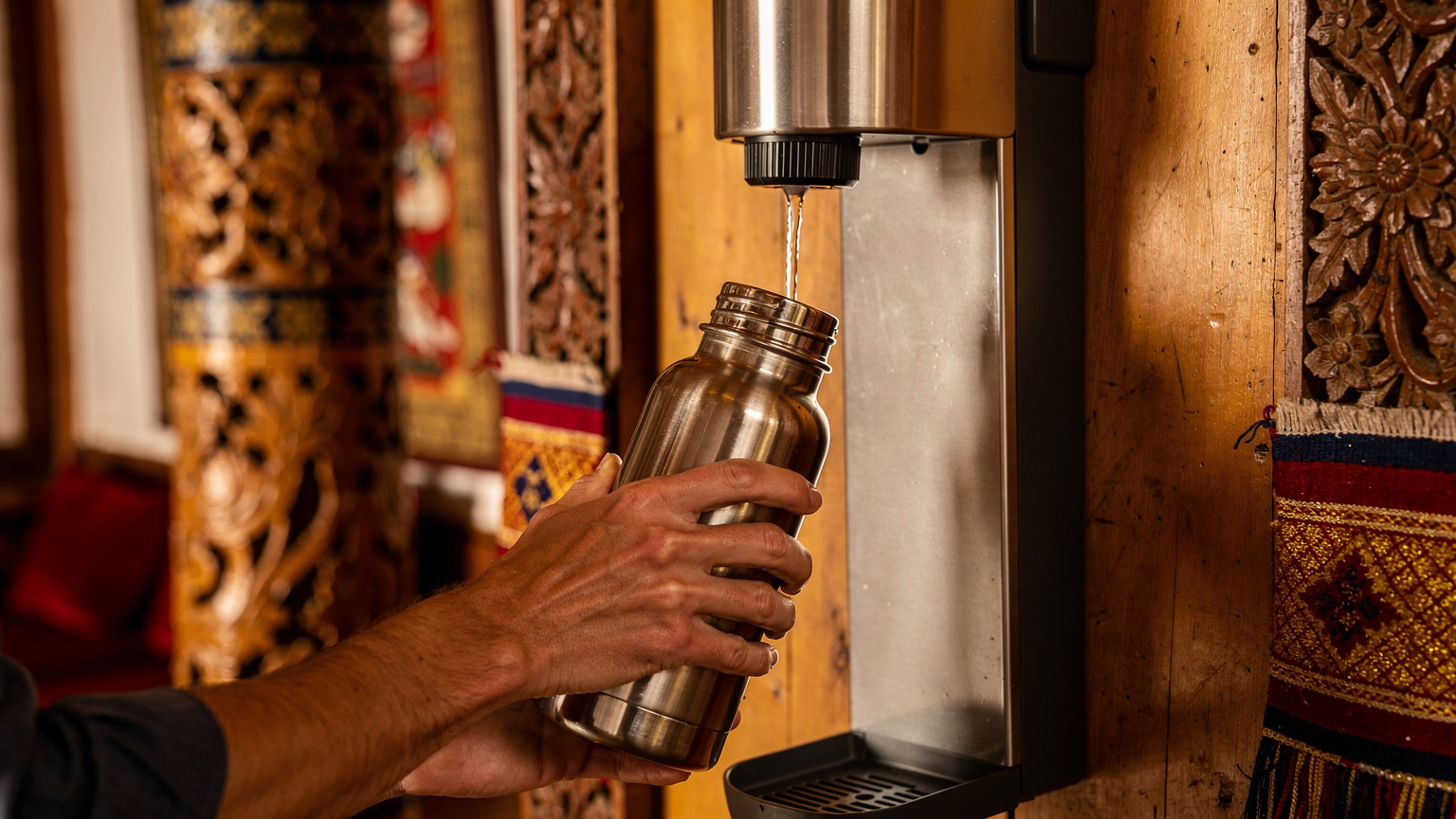 Close-up of a traveler's hands refilling a metal reusable water bottle from a modern water filter dispenser inside a cozy, traditionally decorated Bhutanese guesthouse lobby.