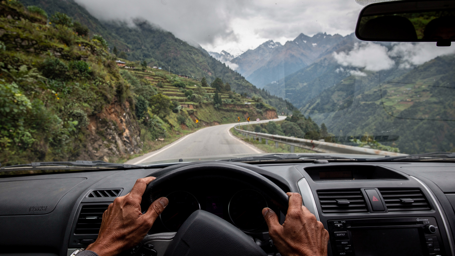 A local Bhutanese driver carefully maneuvering a white SUV along a narrow, winding mountain road, with lush green valleys and distant peaks visible through the windshield on a cloudy day.