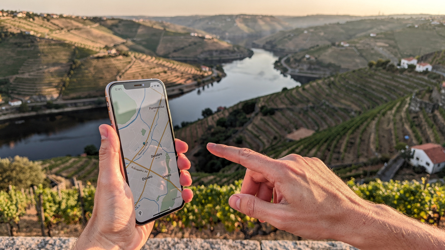 Mão a segurar um iPhone 18 com mapa offline do Douro, apontando para uma vinha distante.