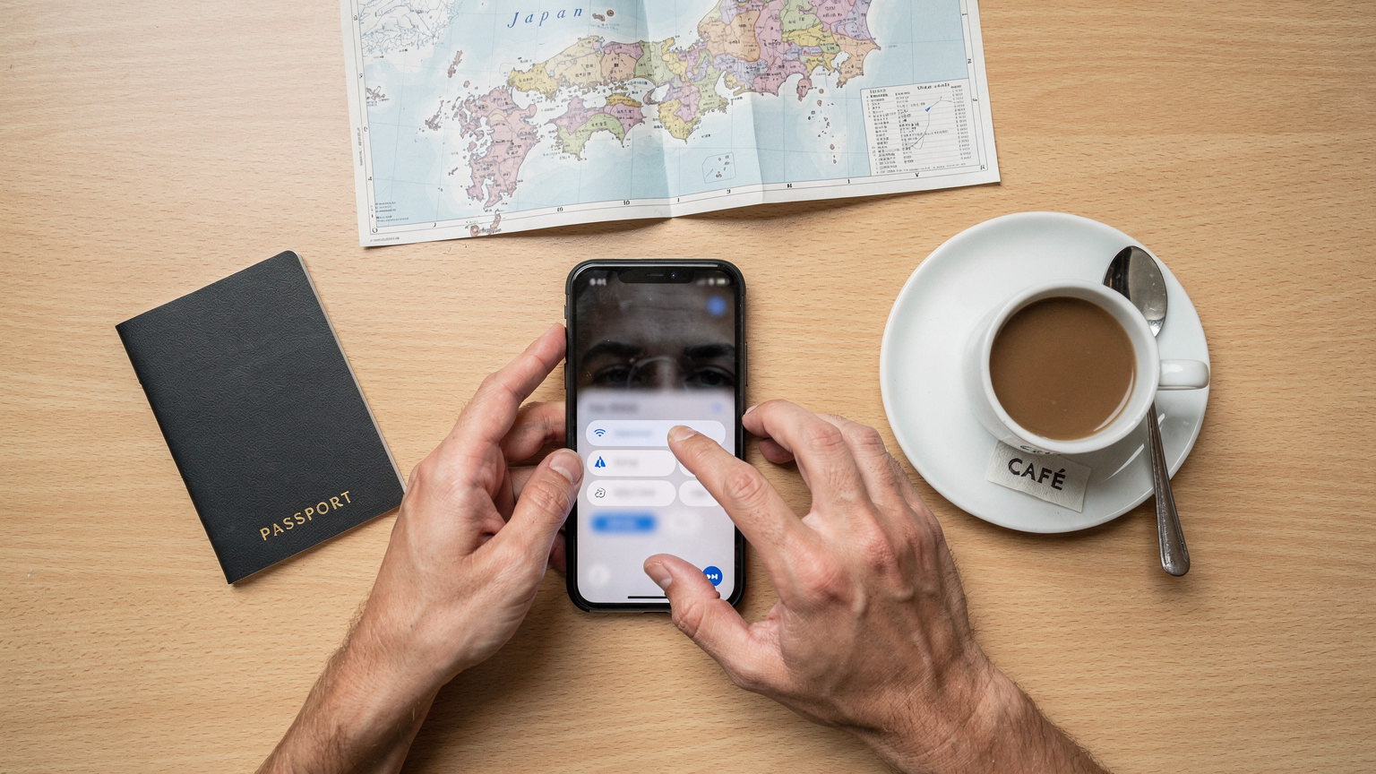 Pandangan overhead dari meja kafe yang rapi, menunjukkan tangan seorang pelancong sedang mengetuk-ngetuk layar ponsel yang menampilkan pengaturan data seluler, di samping paspor, secangkir kopi, dan peta lipat dari Jepang.