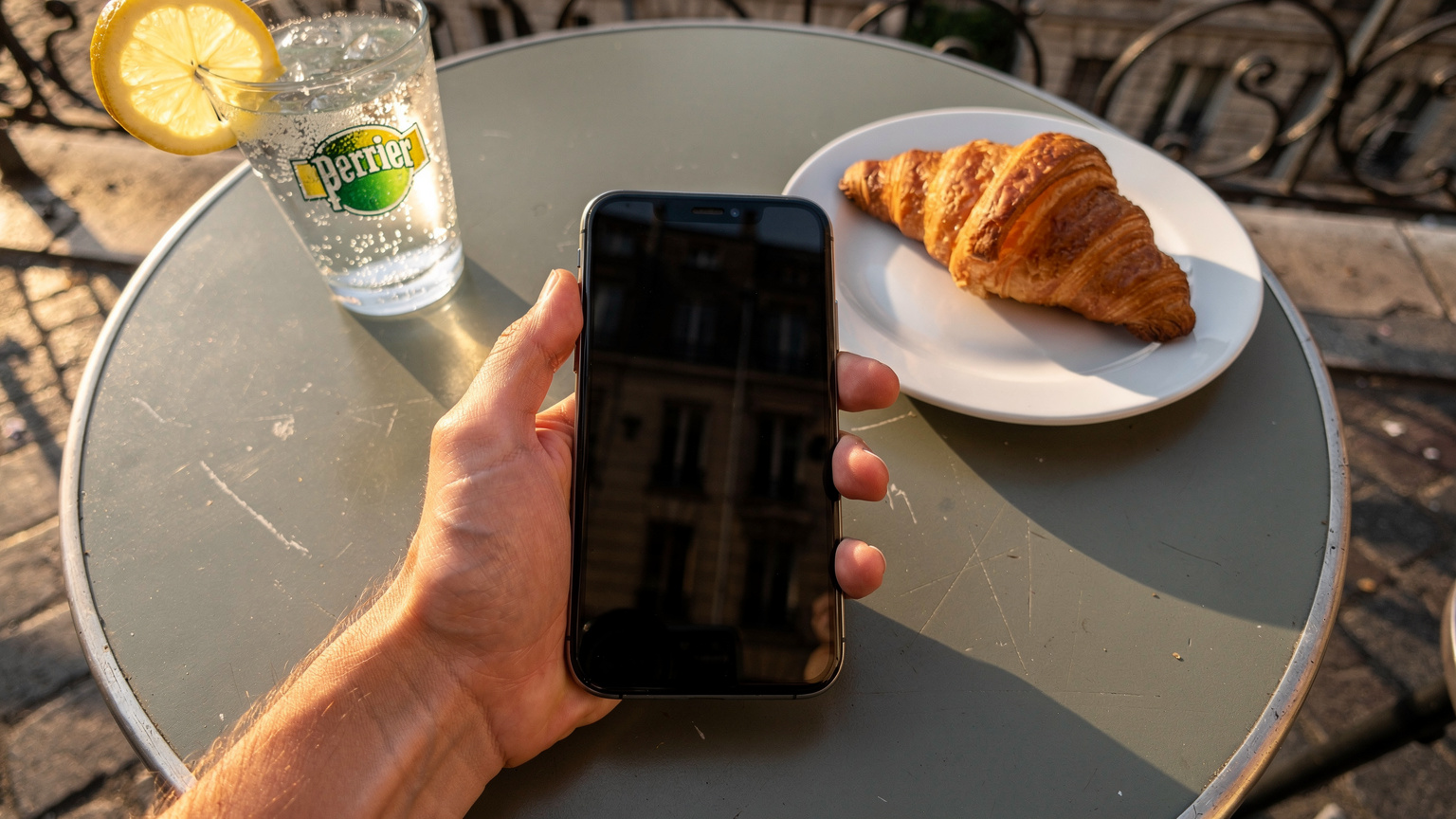 Une main tient un iPhone affichant les réglages de consommation de données cellulaires de l'eSIM Cellesim, posé sur une table de café en plein air à Lyon avec un verre de Perrier et un croissant à côté, par un après-midi ensoleillé.