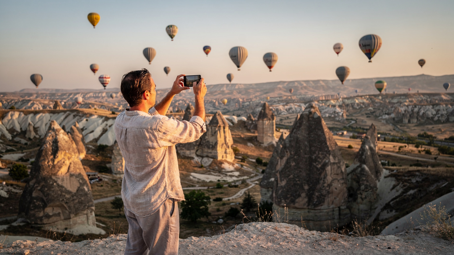 Ein Reisender hält sein Smartphone hoch, um die berühmten Feenkamine in Kappadokien zu fotografieren, während im Hintergrund bei Sonnenaufgang Heißluftballons am Himmel schweben.