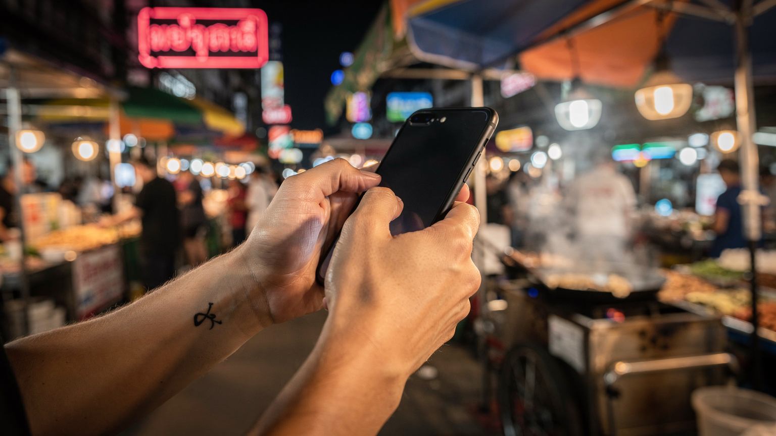 A person's hands holding a smartphone, actively typing or swiping through a messaging app, with a vibrant street food stall in the background in a Bangkok night market, showing cultural details.