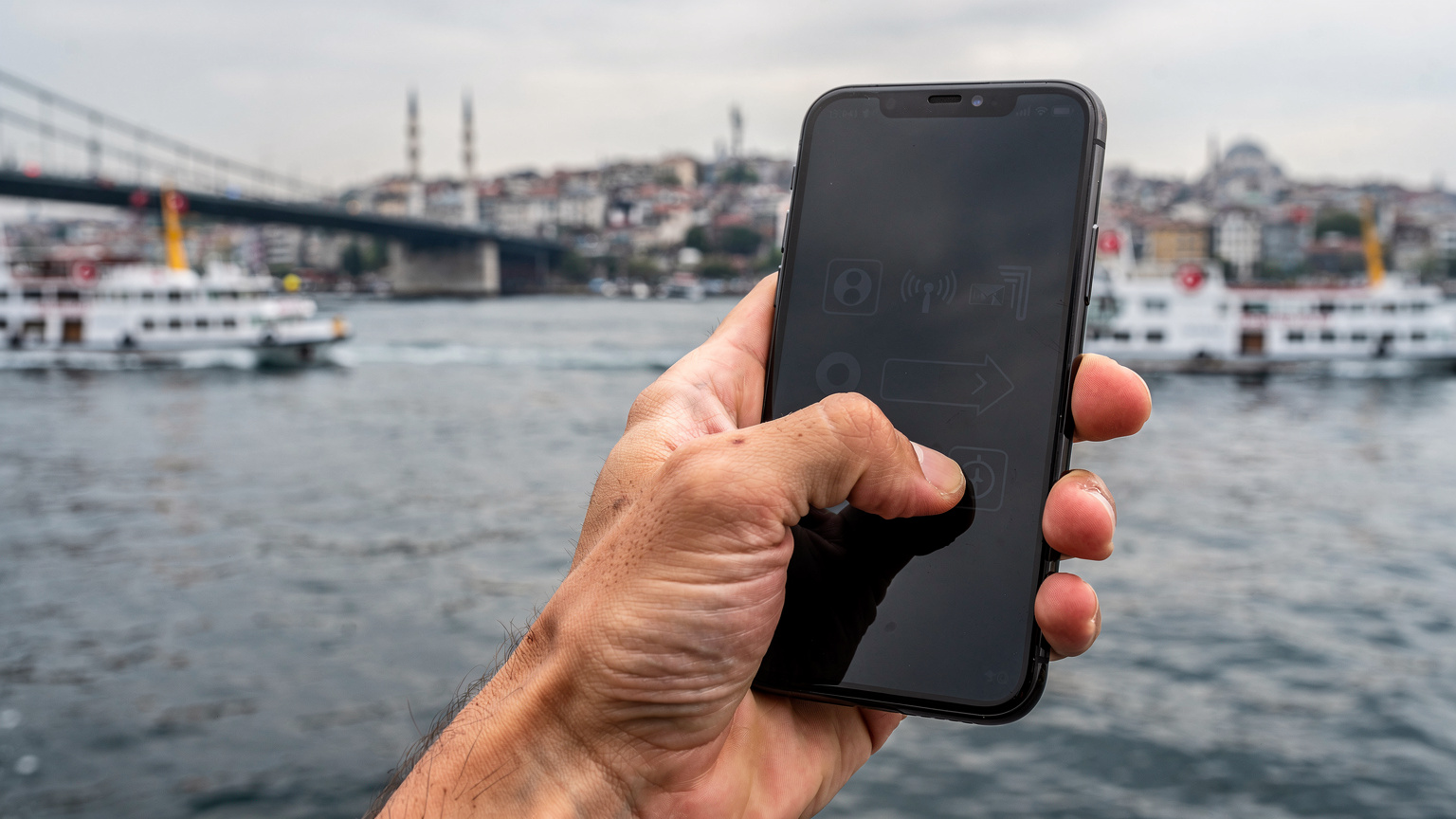 Ένα χέρι κρατά ένα smartphone που δείχνει μήνυμα 'No Service', με το Βόσπορο και την Γέφυρα του Γαλατά στο βάθος, κάτω από ένα συννεφιασμένο ουρανό.