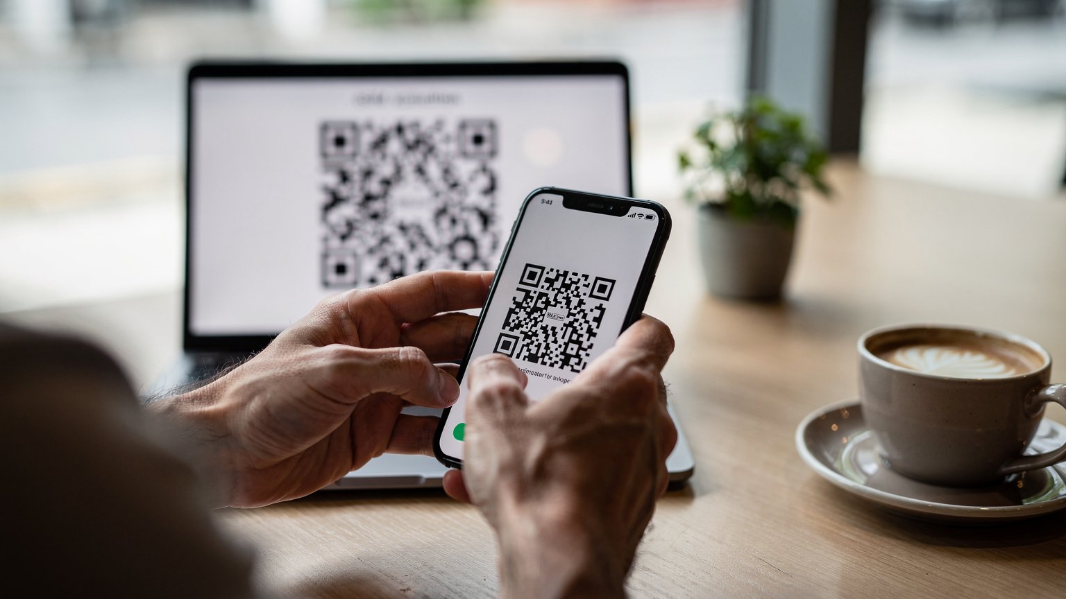 Tangan seorang pengembara memegang telefon pintar, mengimbas kod QR eSIM di skrin komputer riba di sebuah kafe yang tenang di daerah Bugis, Singapura.