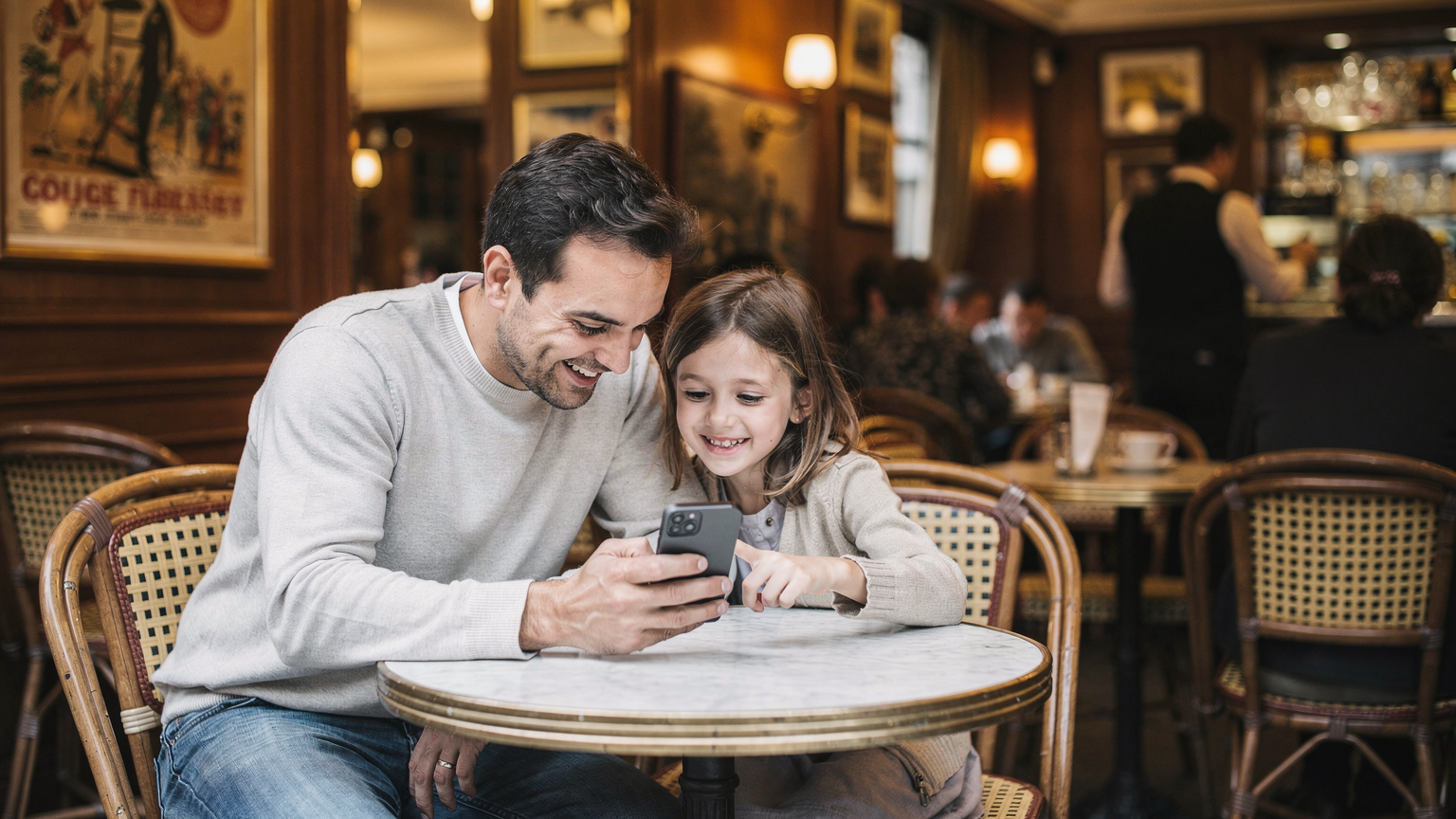 Un père et sa fille regardent attentivement l'écran d'un iPhone, l'enfant montrant du doigt quelque chose qui l'intéresse, dans l'ambiance chaleureuse et animée d'un café parisien.