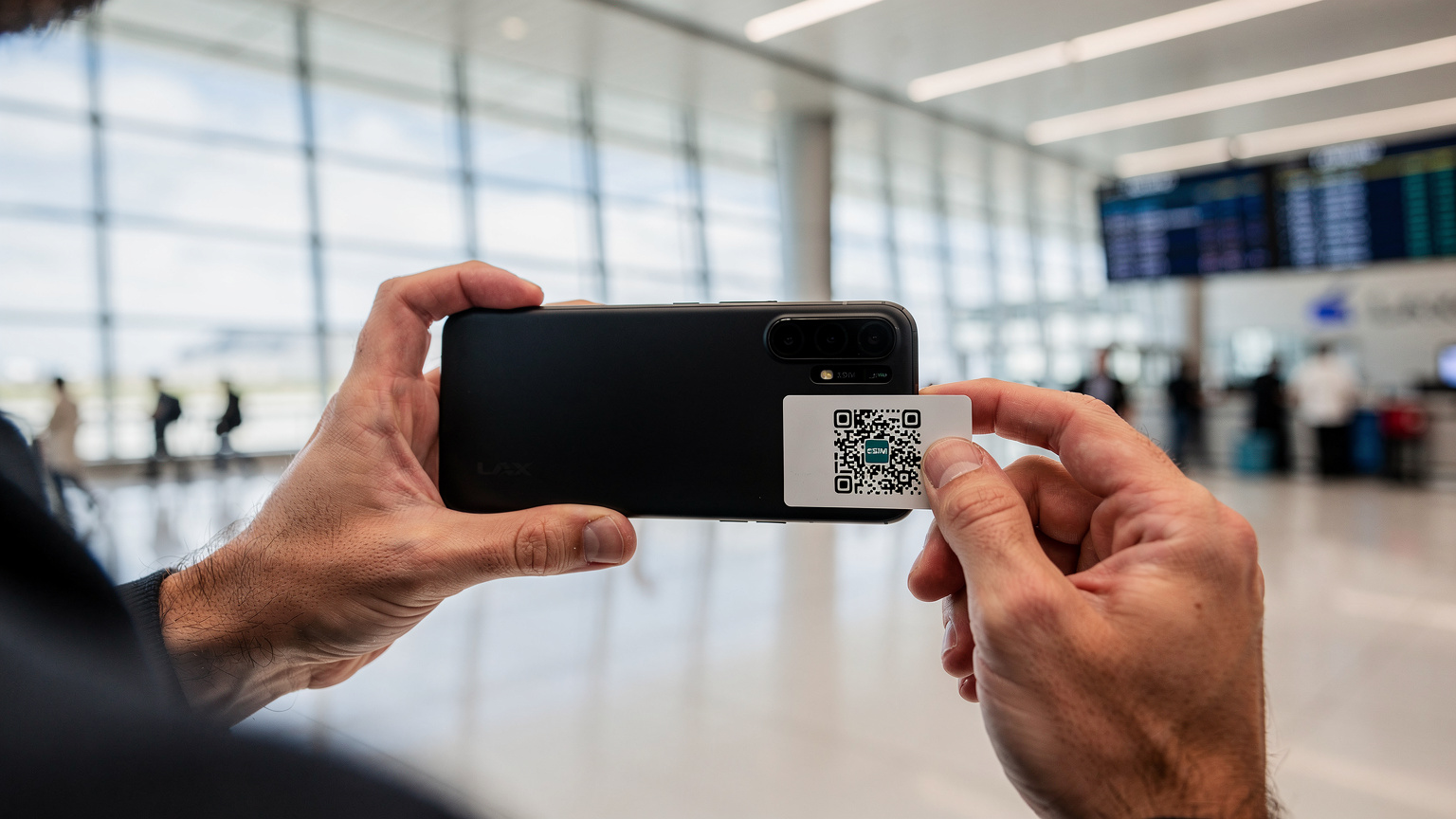 Detailní záběr rukou cestovatele, který v letištní hale na letišti LAX v Los Angeles skenuje QR kód eSIM na svém smartphonu, s digitálními palubními lístky v pozadí.
