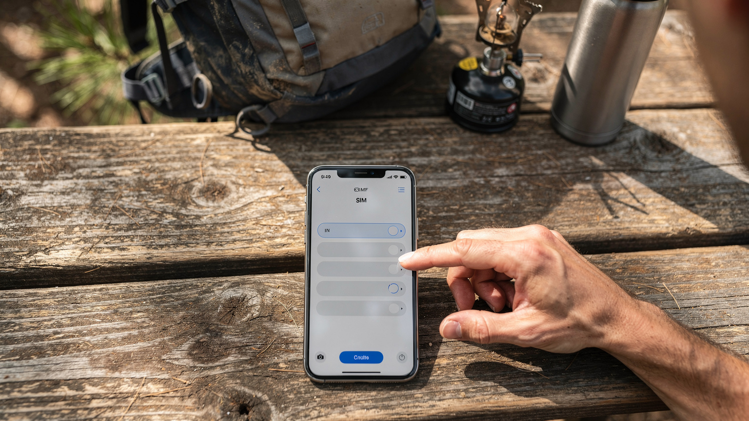Ένα smartphone δείχνει την οθόνη ρυθμίσεων eSIM, με το χέρι ενός ατόμου να επιλέγει ένα προφίλ eSIM, σε ένα σκηνικό με σακίδιο πλάτης και εξοπλισμό κάμπινγκ σε εξωτερικό χώρο.