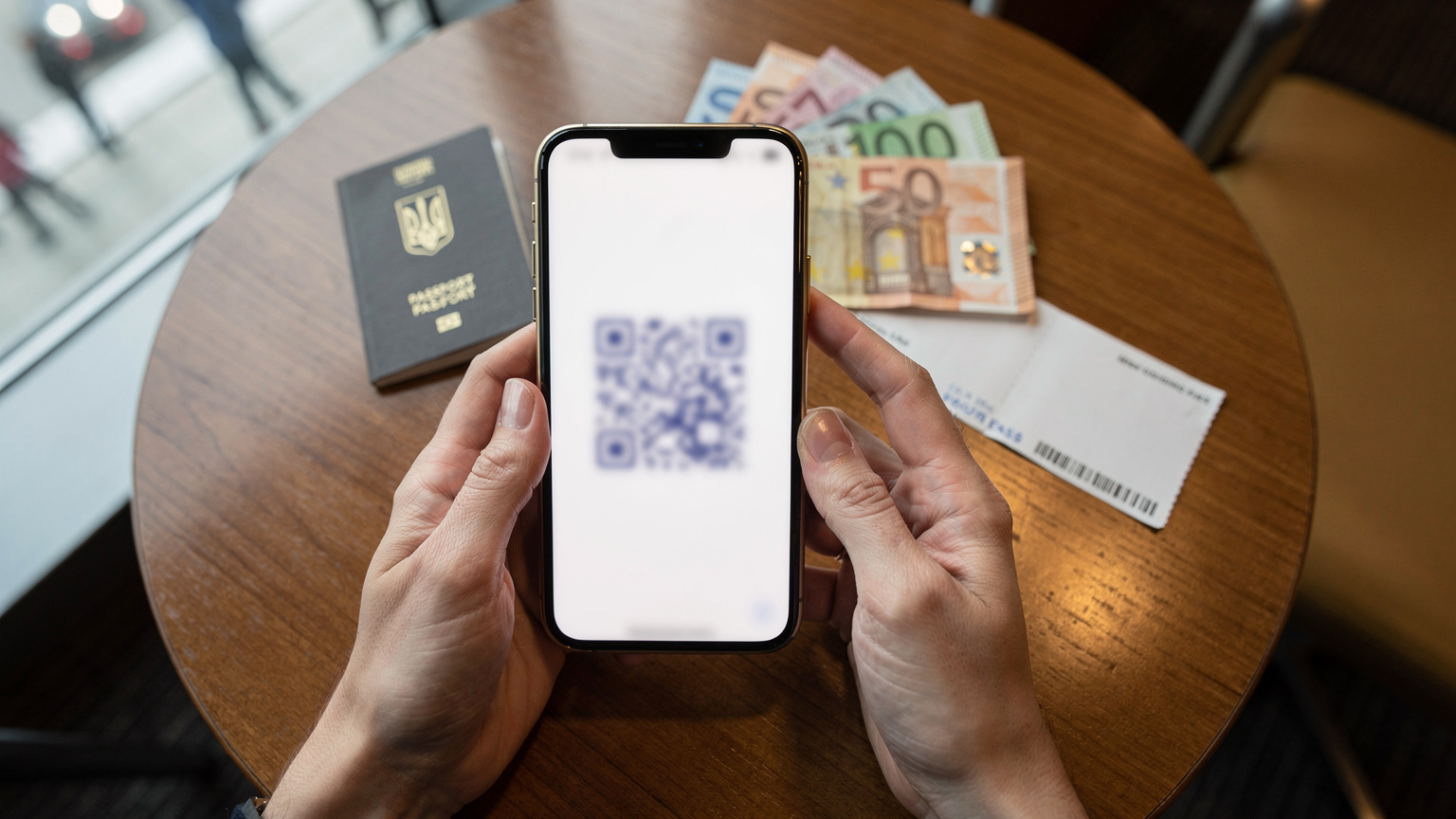 Руки, що тримають смартфон із QR-кодом eSIM на екрані, на тлі паспорта та валюти, що лежать на столі в аеропорту перед вильотом.
