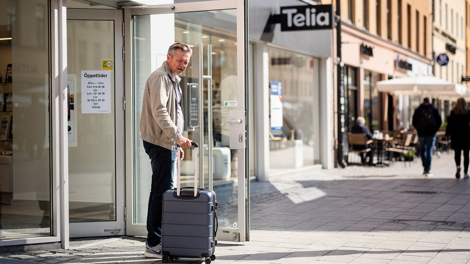 Een toerist staat bij een 'Telia' of 'Telenor' winkel in een drukke winkelstraat in Stockholm, kijkend naar de openingstijden met een lichte uitdrukking van frustratie, een koffer aan zijn zijde.