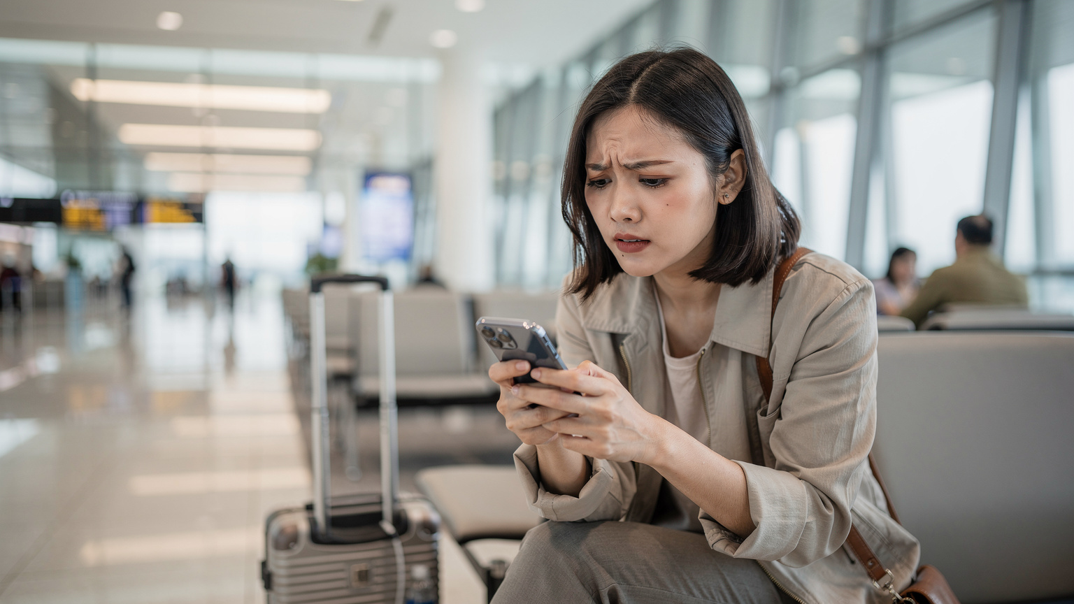 Một phụ nữ Việt Nam trẻ tuổi đang tìm kiếm thông tin về eSIM trên điện thoại của mình với vẻ mặt hơi lo lắng, tại khu vực chờ của sân bay Changi, Singapore, vali hành lý đặt bên cạnh.