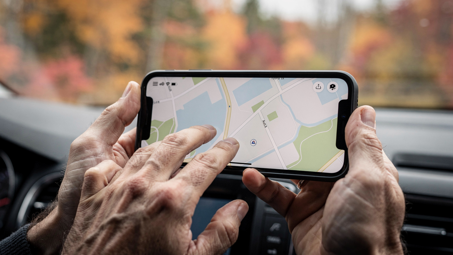 A close-up shot of a parent's hands, with a child's small hand gently resting on one, interacting with a smartphone displaying a map application while driving through a vibrant autumn forest in Quebec, Canada.