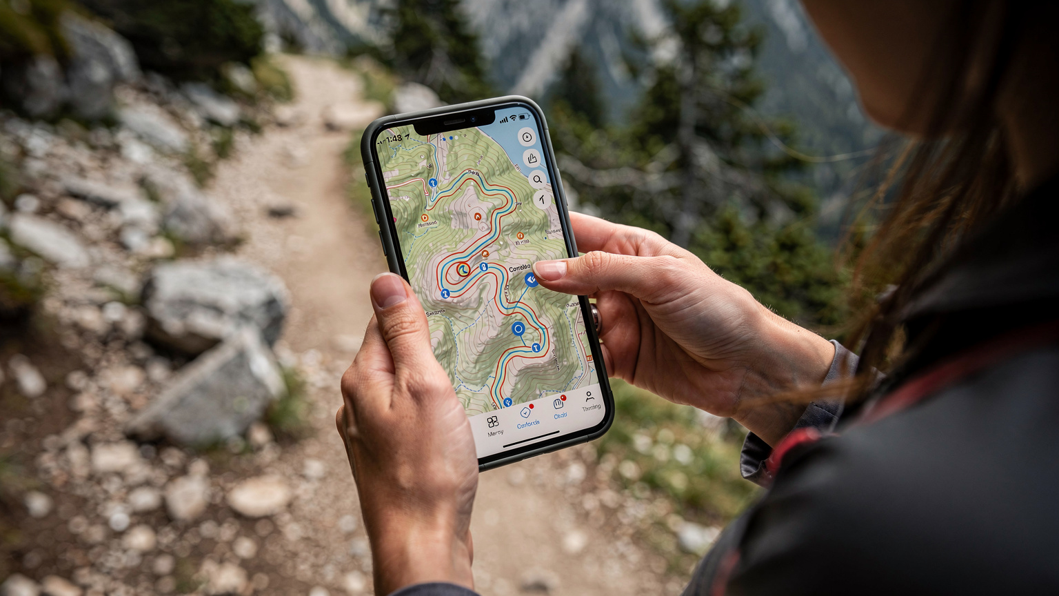 Detailný záber na ruky turistky, ktorá drží smartfón s otvorenou aplikáciou mapy a navigácie, pričom si kontroluje trasu pred turistickým chodníkom v Tatrách.