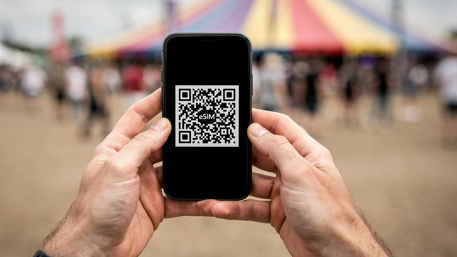 Tæt på-billede af hænder, der holder en smartphone, hvor en QR-kode til eSIM-aktivering vises på skærmen, med sløret festivaltelt og publikum i baggrunden.