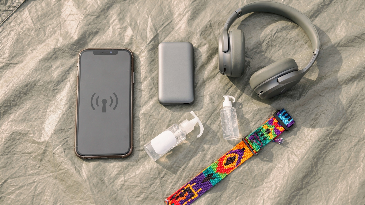 Et organiseret fladlægning af festivalessentials: en smartphone, en powerbank, høretelefoner, et festivalarmbånd og håndsprit på en teltdug.