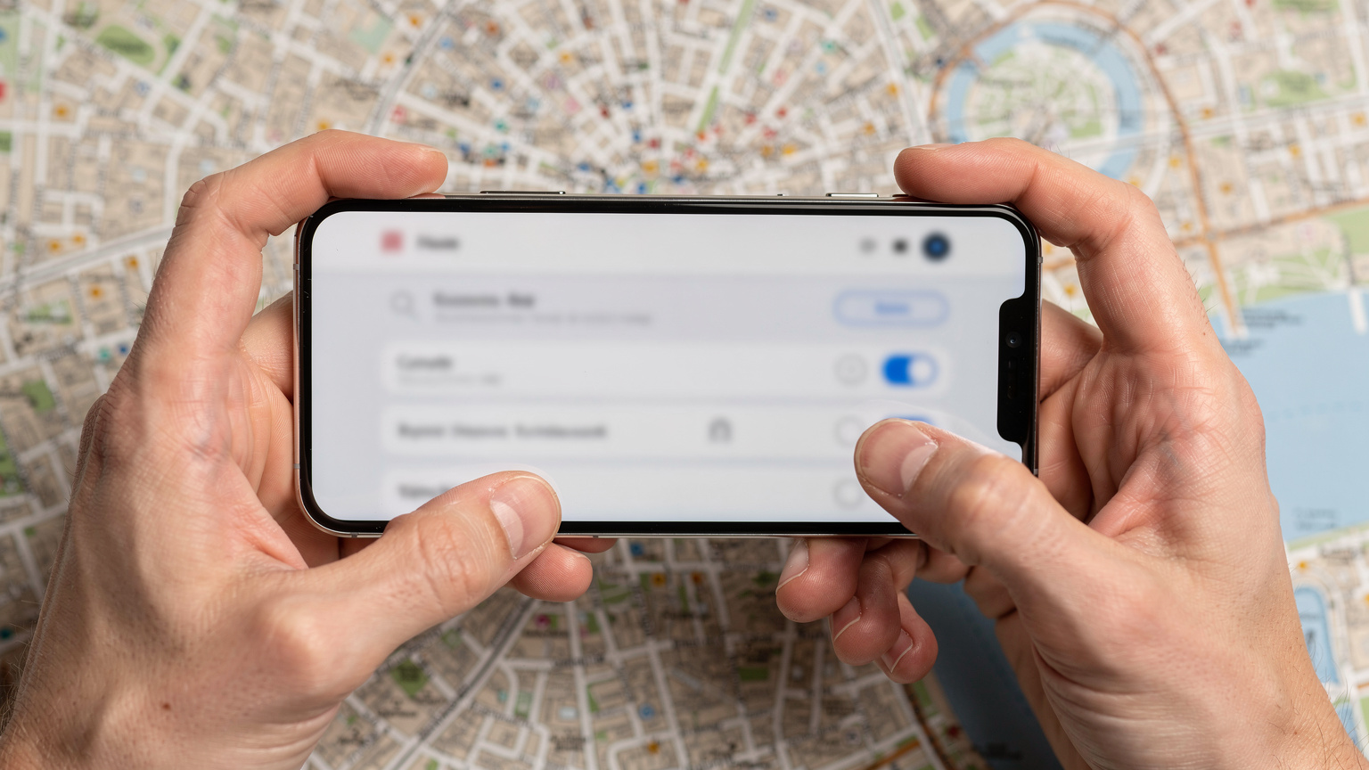 Manos de una persona sosteniendo un smartphone en primer plano, mostrando una interfaz de ajustes de conectividad sin texto legible, con un mapa digital desenfocado de Londres de fondo en la pantalla.