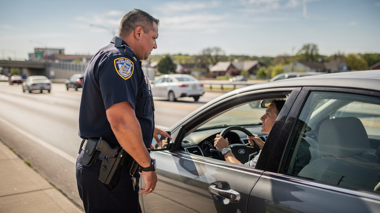 Un ofițer de poliție american, în uniformă, vorbește cu un șofer printr-o fereastră deschisă a mașinii, pe o autostradă, în timpul unei opriri de rutină.