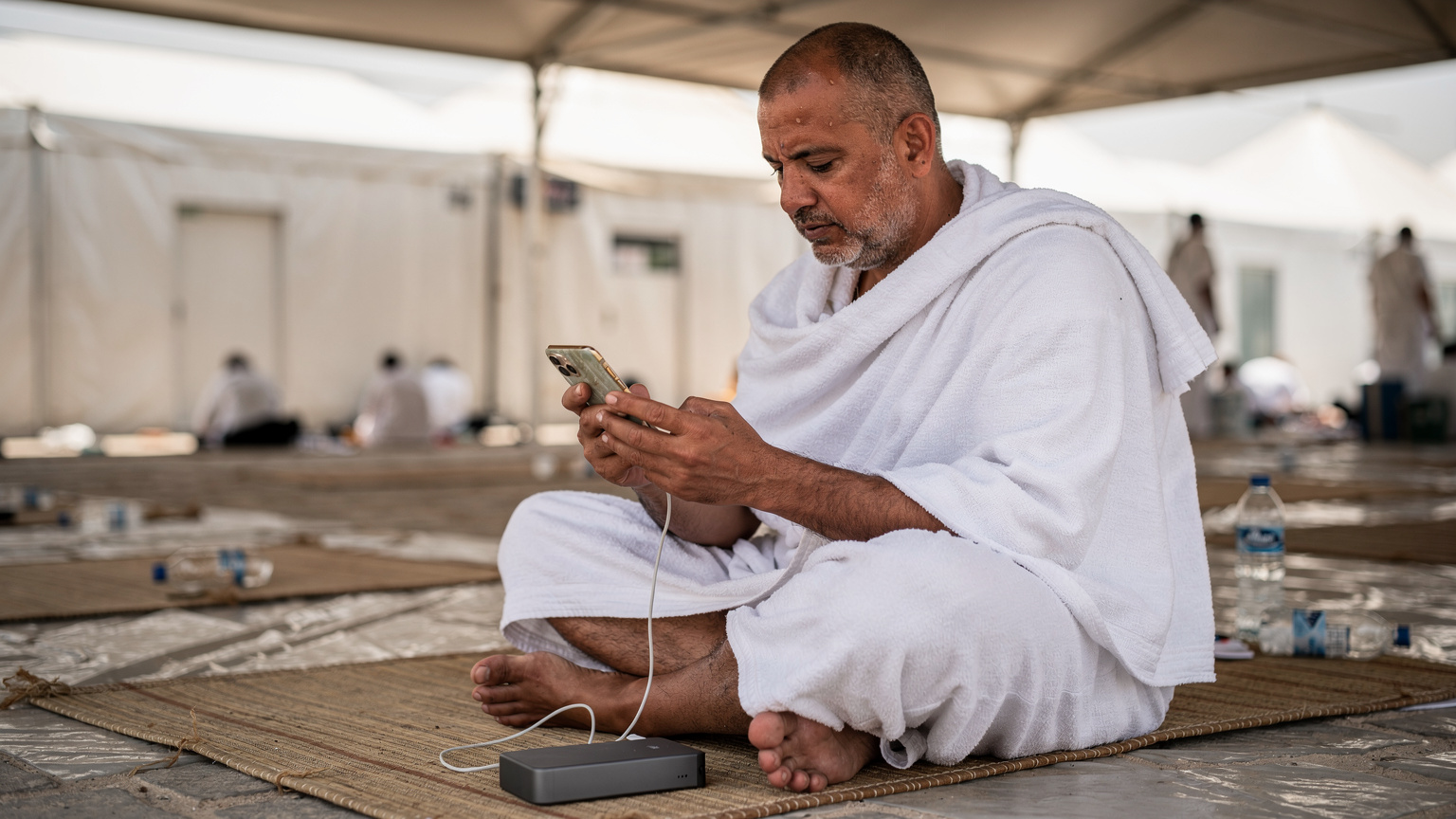 Seorang jemaah haji lelaki sedang mengecas telefon pintarnya menggunakan power bank di kawasan rehat yang teduh di Mina, semasa musim Haji.