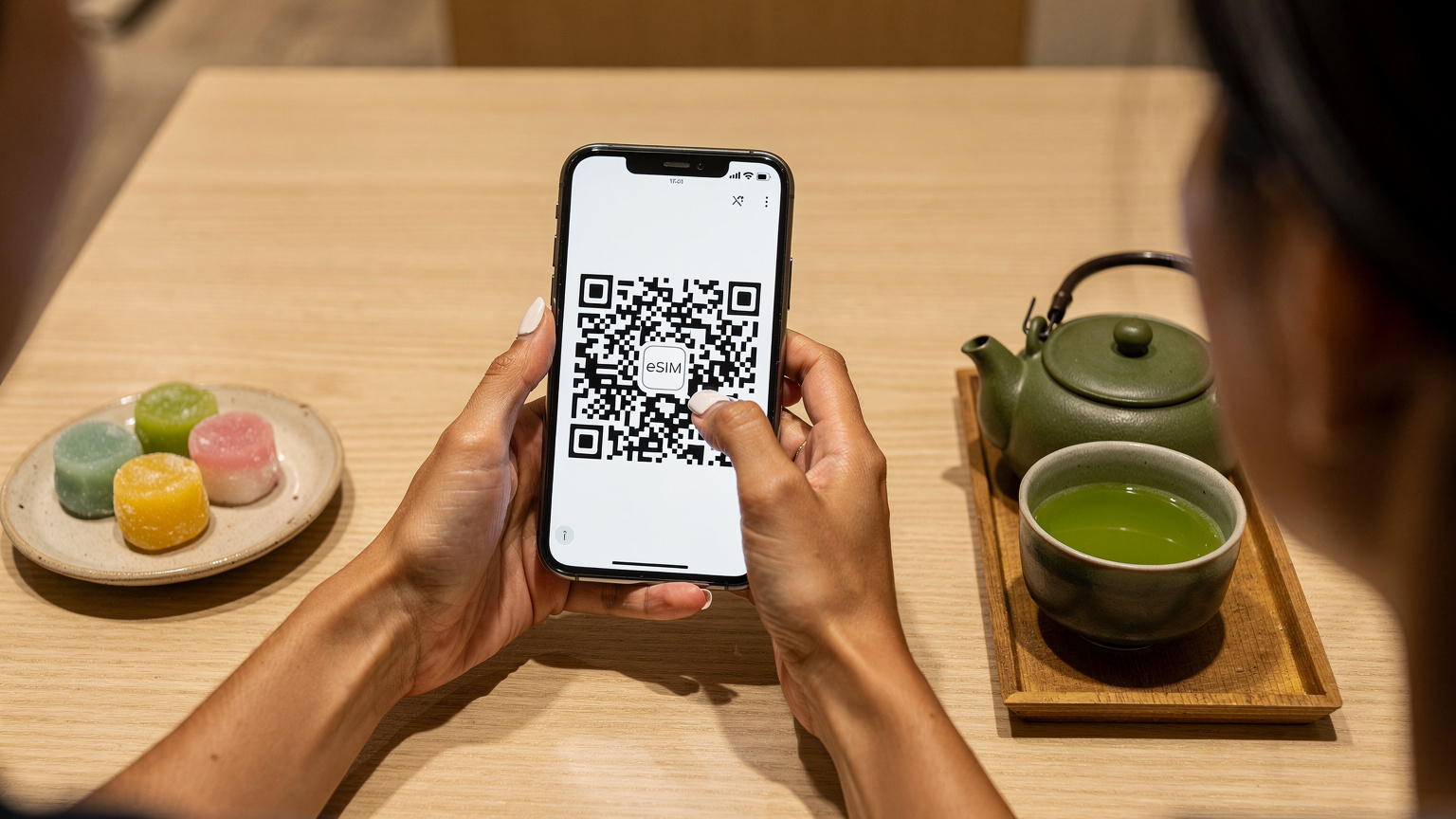 Bir Türk gezgin, Tokyo'daki bir kafede otururken, akıllı telefonunda QR kod tarayarak eSIM aktivasyonunu yapıyor. Masasında Japon yeşil çayı ve küçük bir mochi tatlısı var.