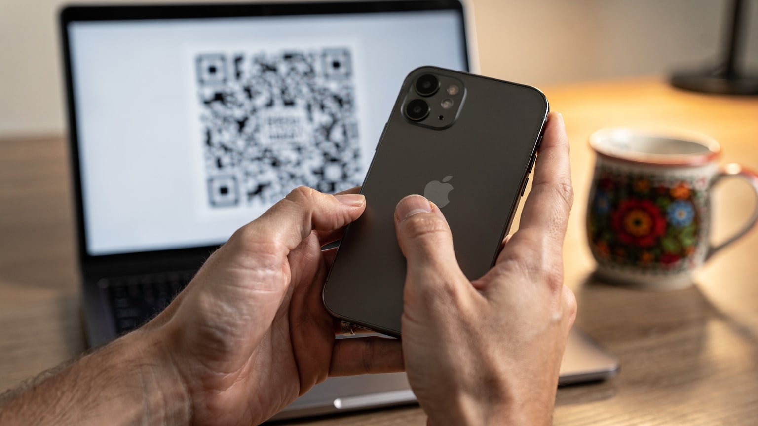 Zbliżenie na dłonie osoby aktywującej eSIM na iPhone'ie 18, z kodem QR na ekranie laptopa w tle.