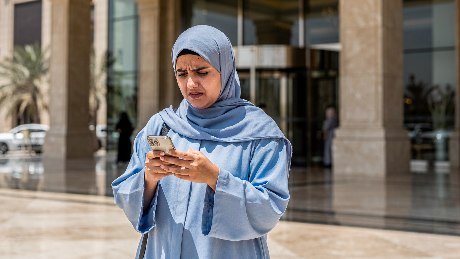 A woman in a hijab looking at her smartphone with a frustrated expression, standing in front of the ornate entrance of a large building in Saudi Arabia.