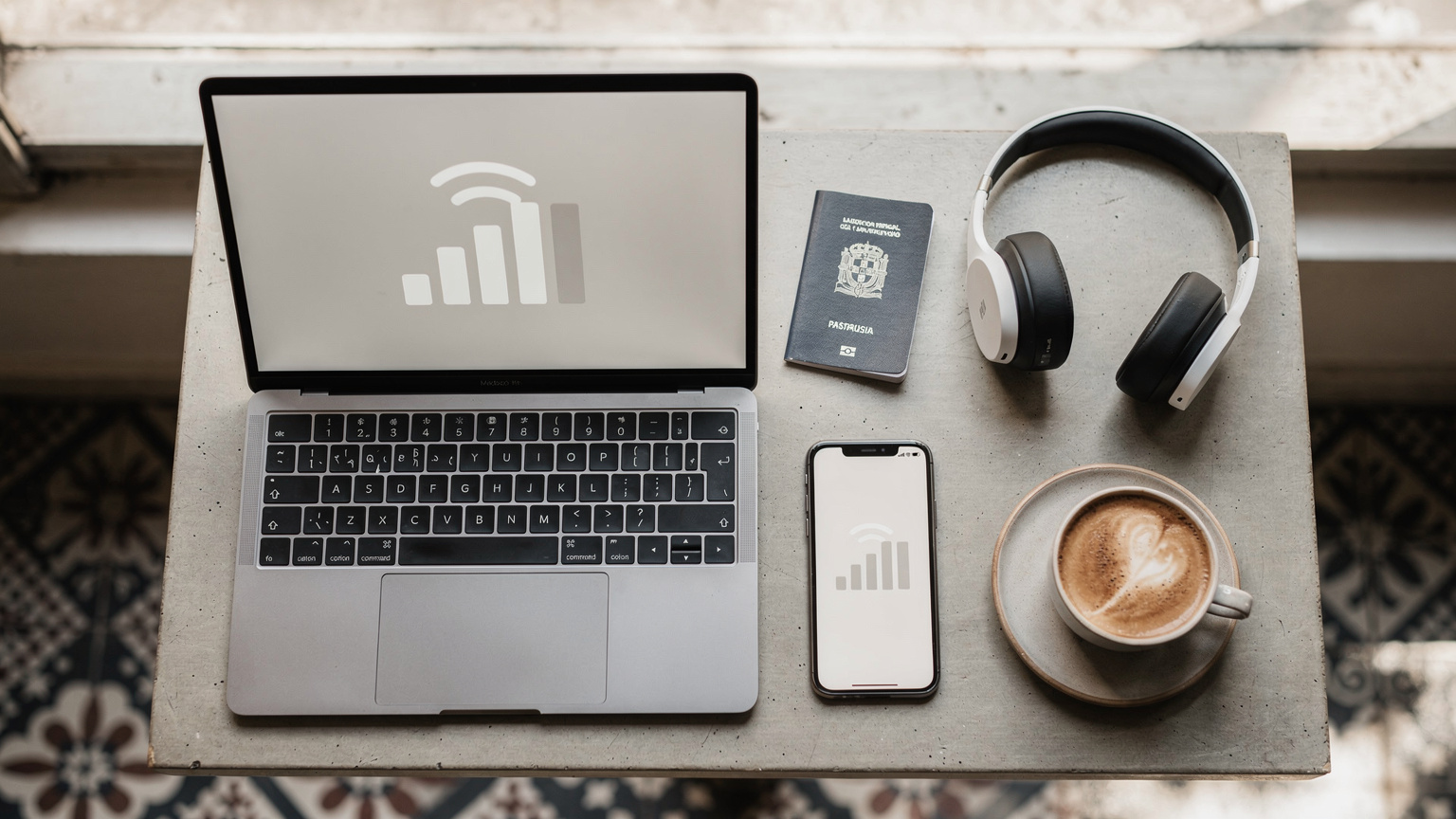 Uma mesa de café com um laptop aberto, um smartphone com um gráfico de barras de conectividade forte, um passaporte e uma xícara de café, em um ambiente de coworking moderno no bairro de Campo de Ourique, em Lisboa.