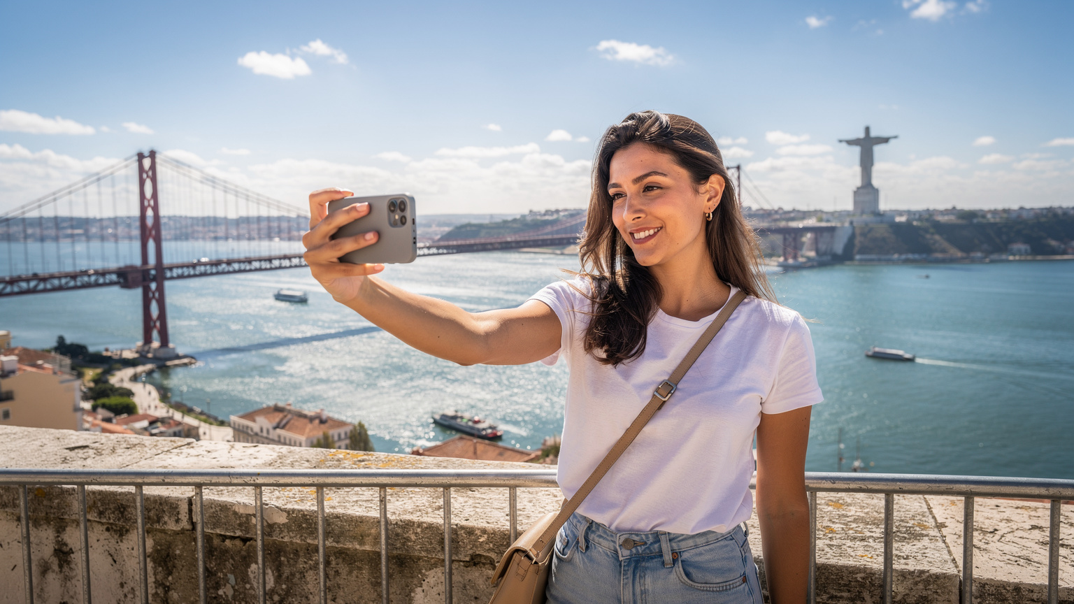 Uma jovem com um smartphone na mão, sorrindo e tirando uma selfie com a Ponte 25 de Abril e o Cristo Rei ao fundo, em um dia ensolarado em Lisboa. Ela está conectada, vivendo o momento.
