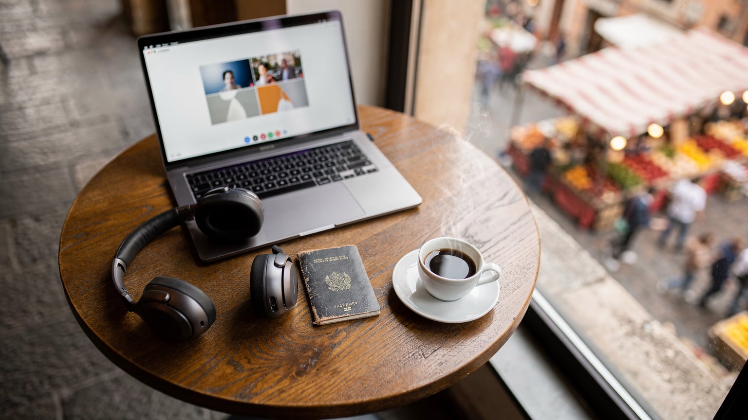 Un tavolino con un laptop aperto su una videochiamata, un passaporto, un paio di cuffie e una tazza di caffè, con una finestra che offre una vista sfocata di un mercato tradizionale italiano.