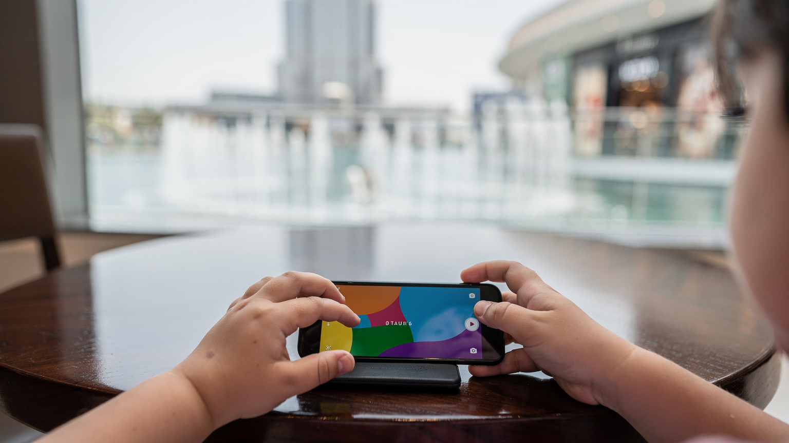 Крупный план рук ребенка, играющего в мобильную игру на смартфоне, лежащем на полированном столике кафе в торговом центре Дубая, с видом на танцующие фонтаны.
