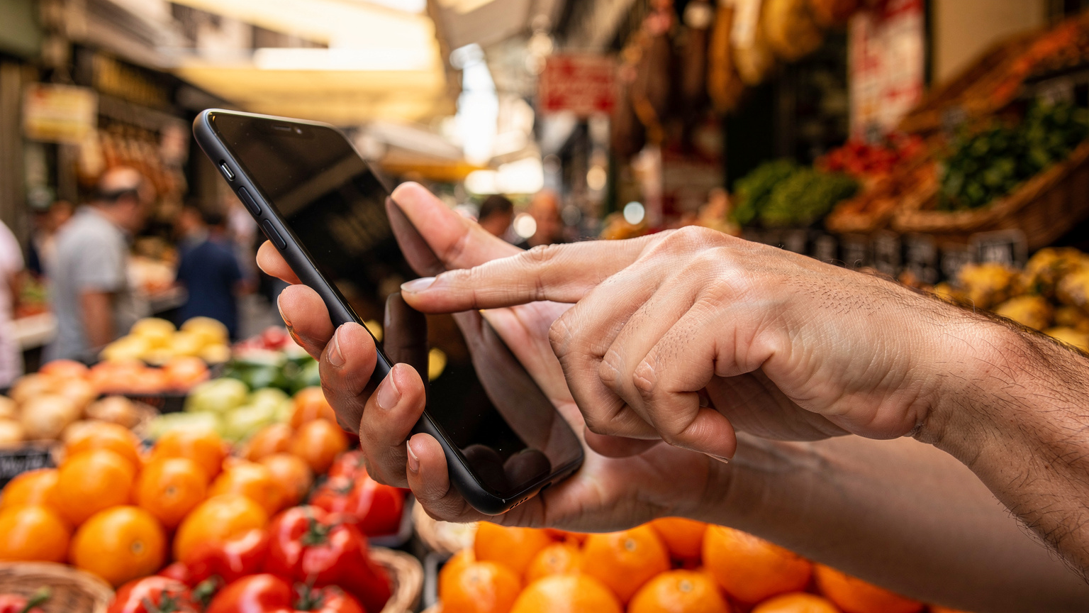 Een close-up van handen die een smartphone vasthouden, waarbij de focus ligt op de vingers die over het scherm vegen, met op de achtergrond de levendige kleuren van een Spaanse markt.