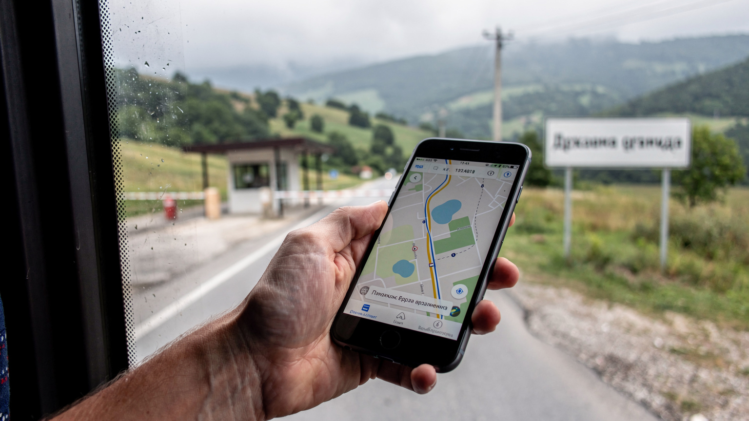 Bir otobüs penceresinden görünen, yeşil tepelerin arasından geçen bir sınır kapısı. Pencerenin önünde, bir gezginin elinde açık bir harita uygulaması olan telefon görünüyor, internet bağlantısı sorunsuz çalışıyor.