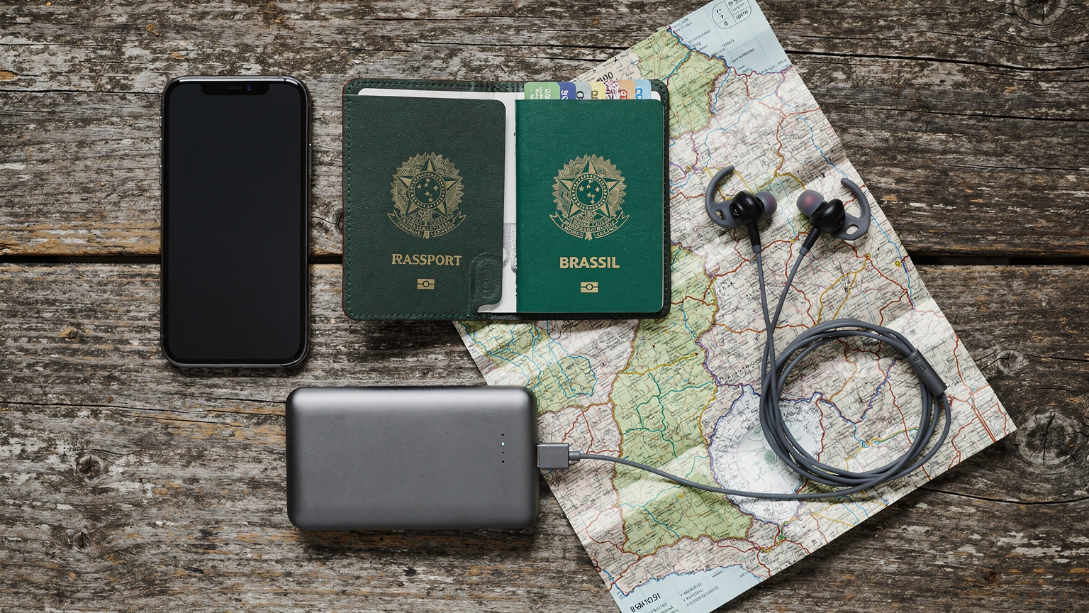 Uma visão aérea de um iPhone, um power bank robusto, fones de ouvido intra-auriculares, um mapa de papel levemente amassado e um passaporte brasileiro aberto, dispostos sobre uma superfície de madeira rústica, simbolizando a preparação para uma viagem.