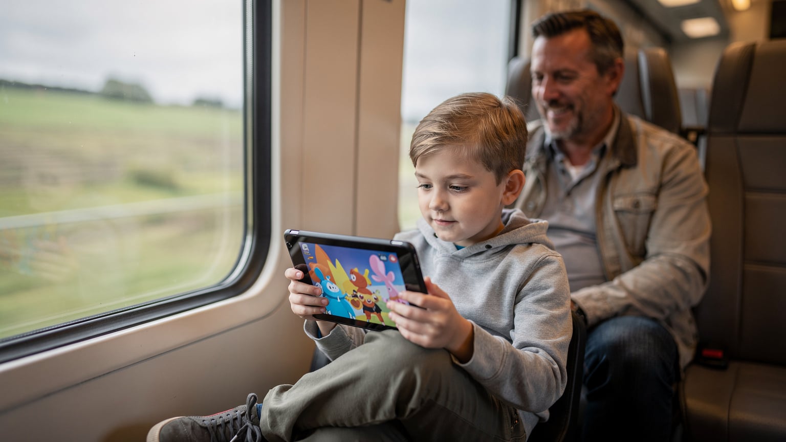 A child intently watching a cartoon on a tablet during a long train journey, with a parent looking on with a relieved smile in the background.
