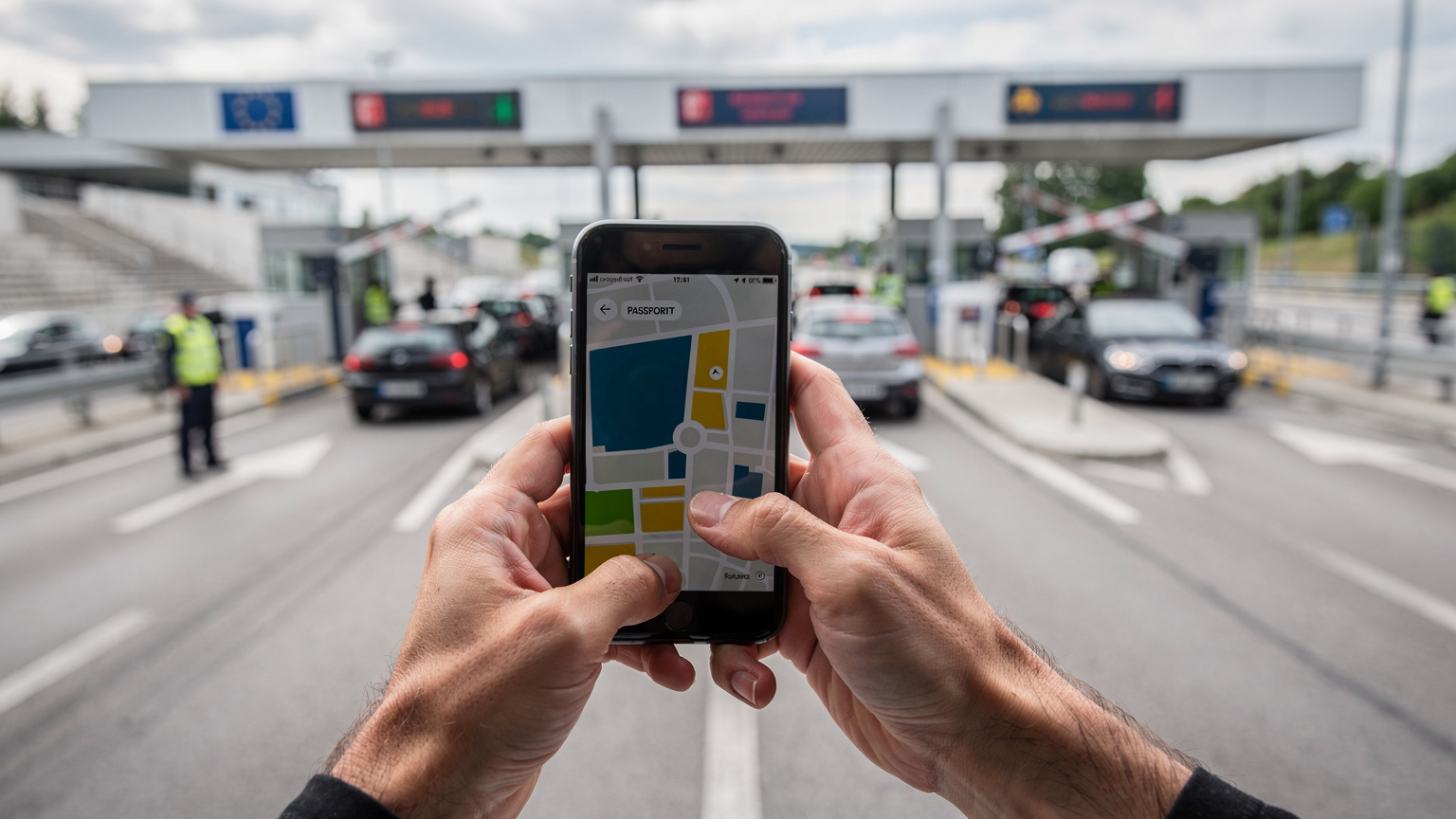 Χέρια που κρατούν ένα smartphone, δείχνοντας την οθόνη με χάρτες, ενώ διασχίζουν ένα πολυσύχναστο συνοριακό σημείο ελέγχου στην Ευρώπη.