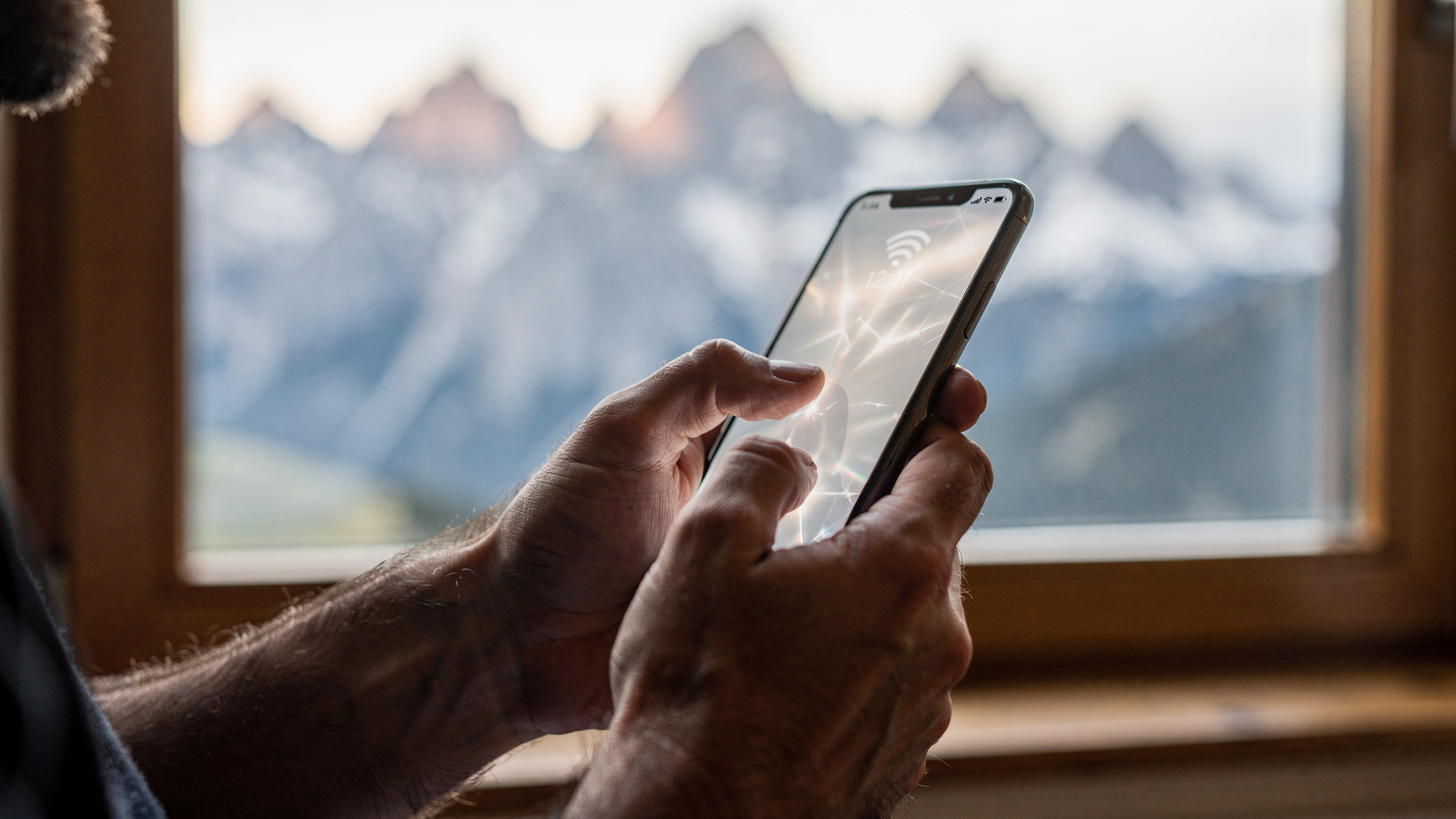 Detailní záběr rukou digitálního nomáda, který drží chytrý telefon a sleduje ukazatel síly signálu, zatímco sedí u okna s výhledem na slovenské hory. Telefon ukazuje síť 4G/5G.