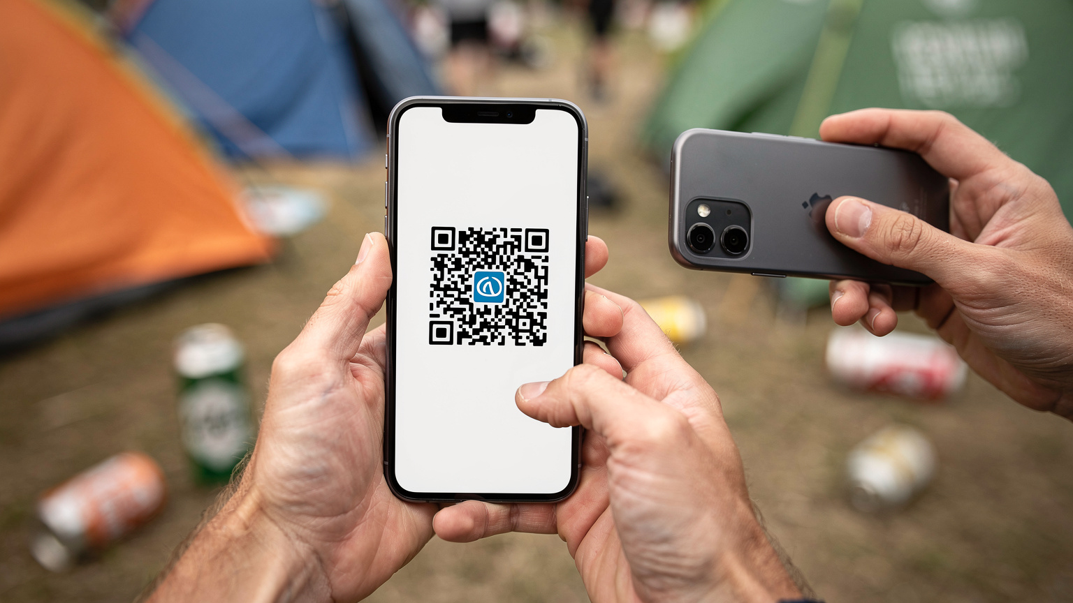 Et tæt-op billede af to hænder, der holder en smartphone, hvor en Cellesim QR-kode er tydeligt synlig på skærmen, i færd med at blive scannet af en anden telefon. Baggrunden er sløret med campingudstyr og festivalstemning.