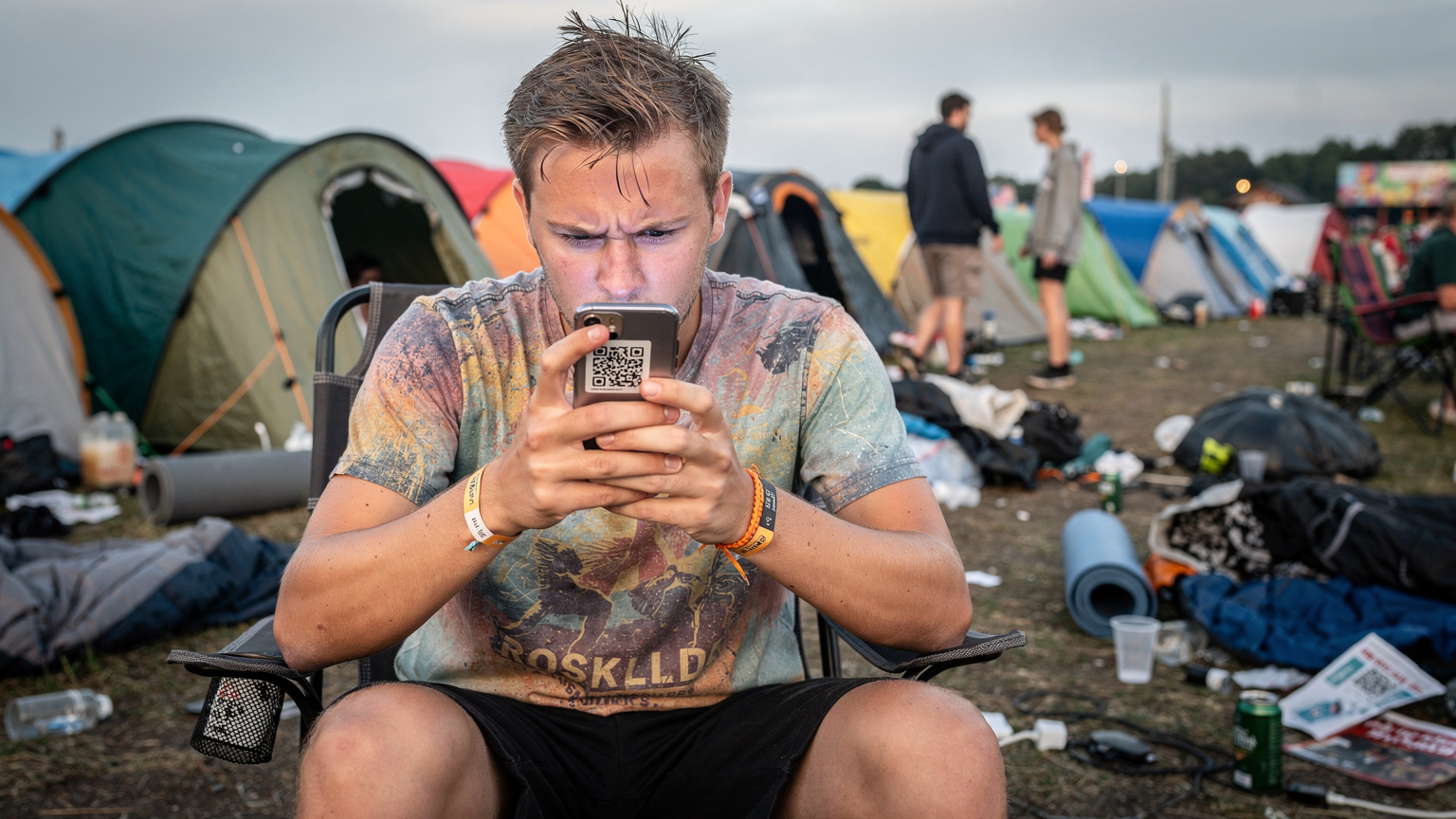 En ung mand med festivalarmbånd og en farverig T-shirt sidder på en sammenklappelig campingstol og holder sin smartphone tæt på ansigtet, mens han febrilsk forsøger at scanne en QR-kode for at aktivere sit eSIM på Roskilde Festivals campingområde.
