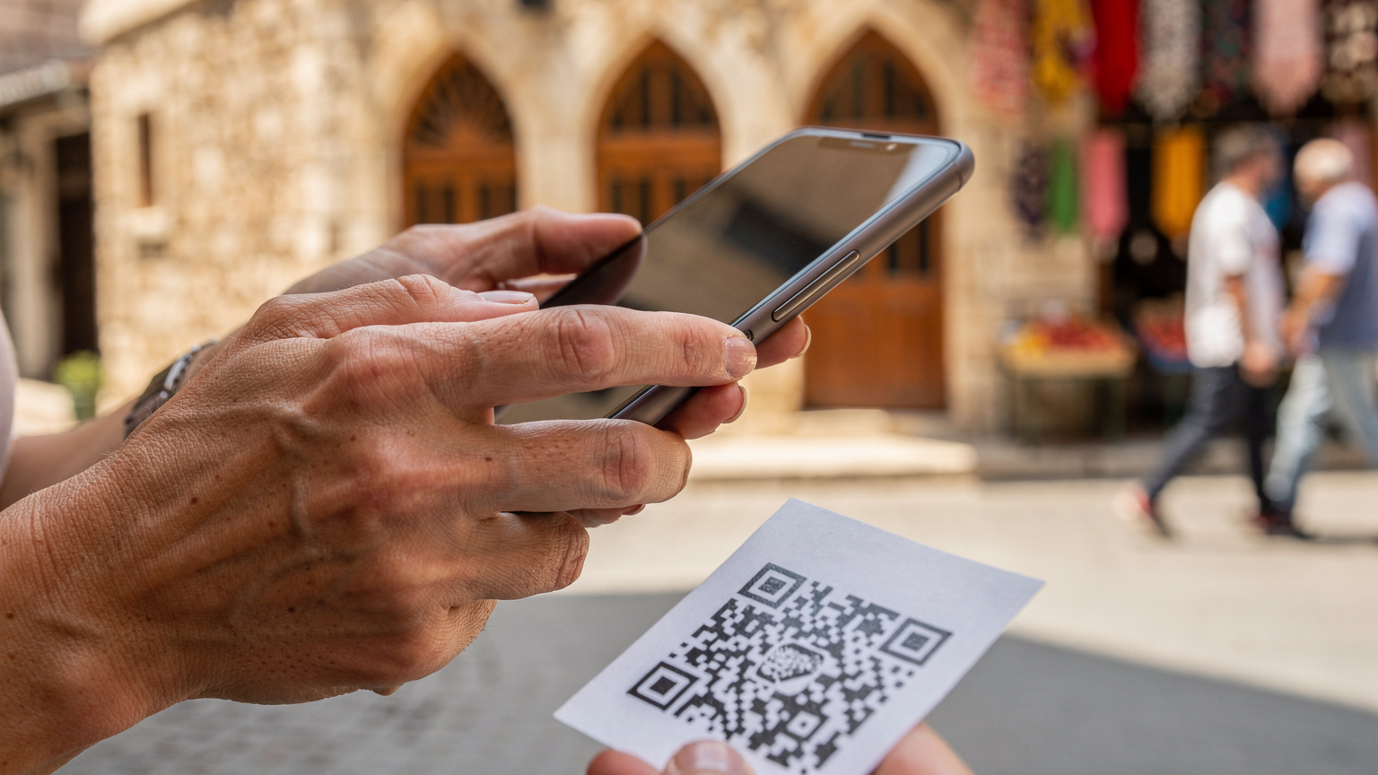 Detailní záběr rukou rodiče, který drží chytrý telefon a skenuje QR kód pro aktivaci eSIM. V pozadí je rozmazaná rušná turecká ulice s historickými budovami a místními lidmi.