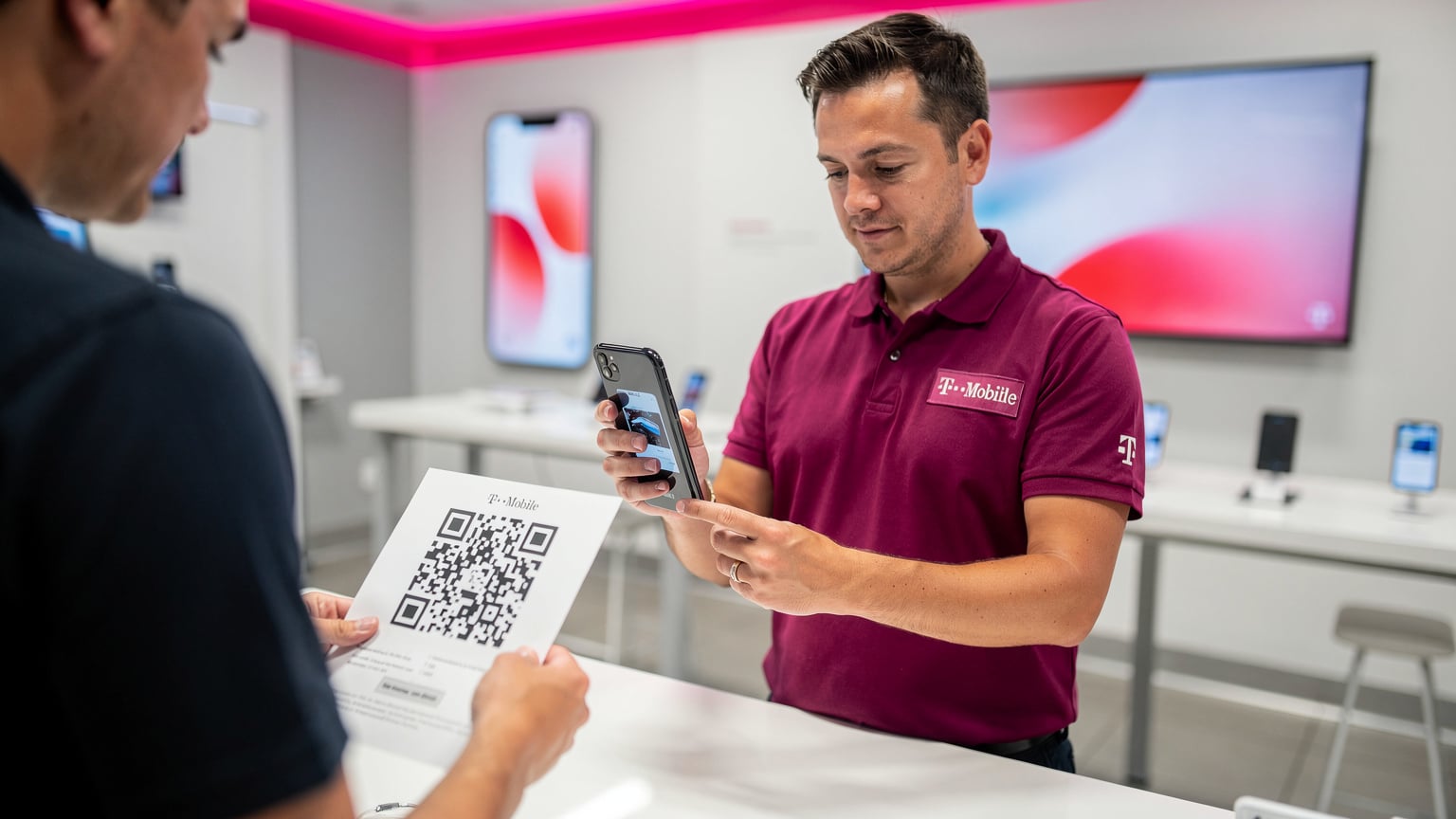 A T-Mobile store employee in a branded shirt is assisting a customer by scanning a QR code on a document with a smartphone, demonstrating the eSIM activation process in a bright, modern retail environment.
