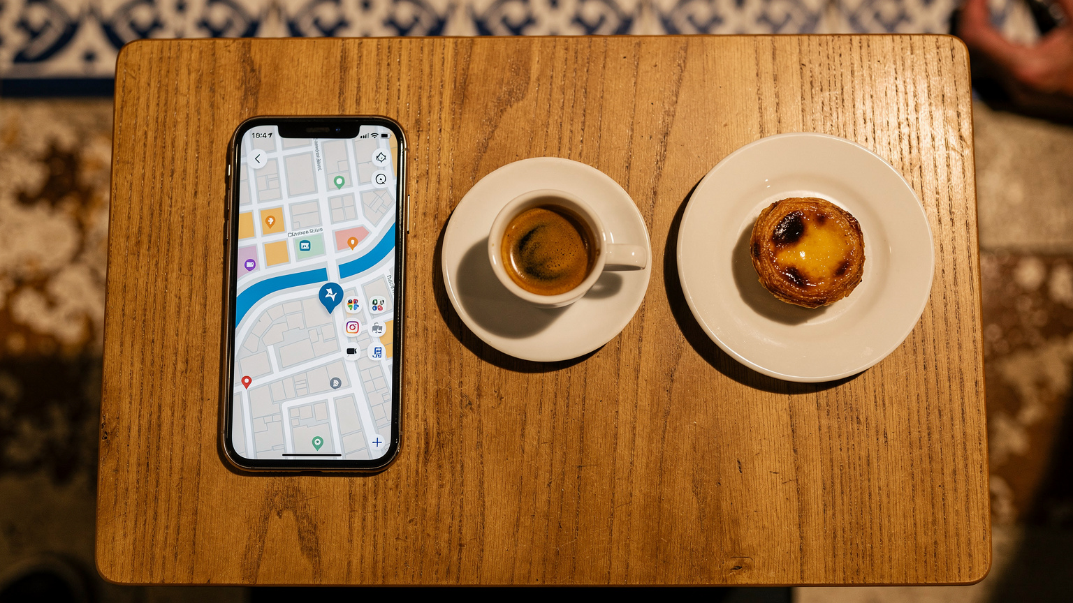 Uma mesa de café em Porto com um iPhone 18 exibindo um mapa, um café bica e um pastel de nata, evocando a experiência de um nômade digital.