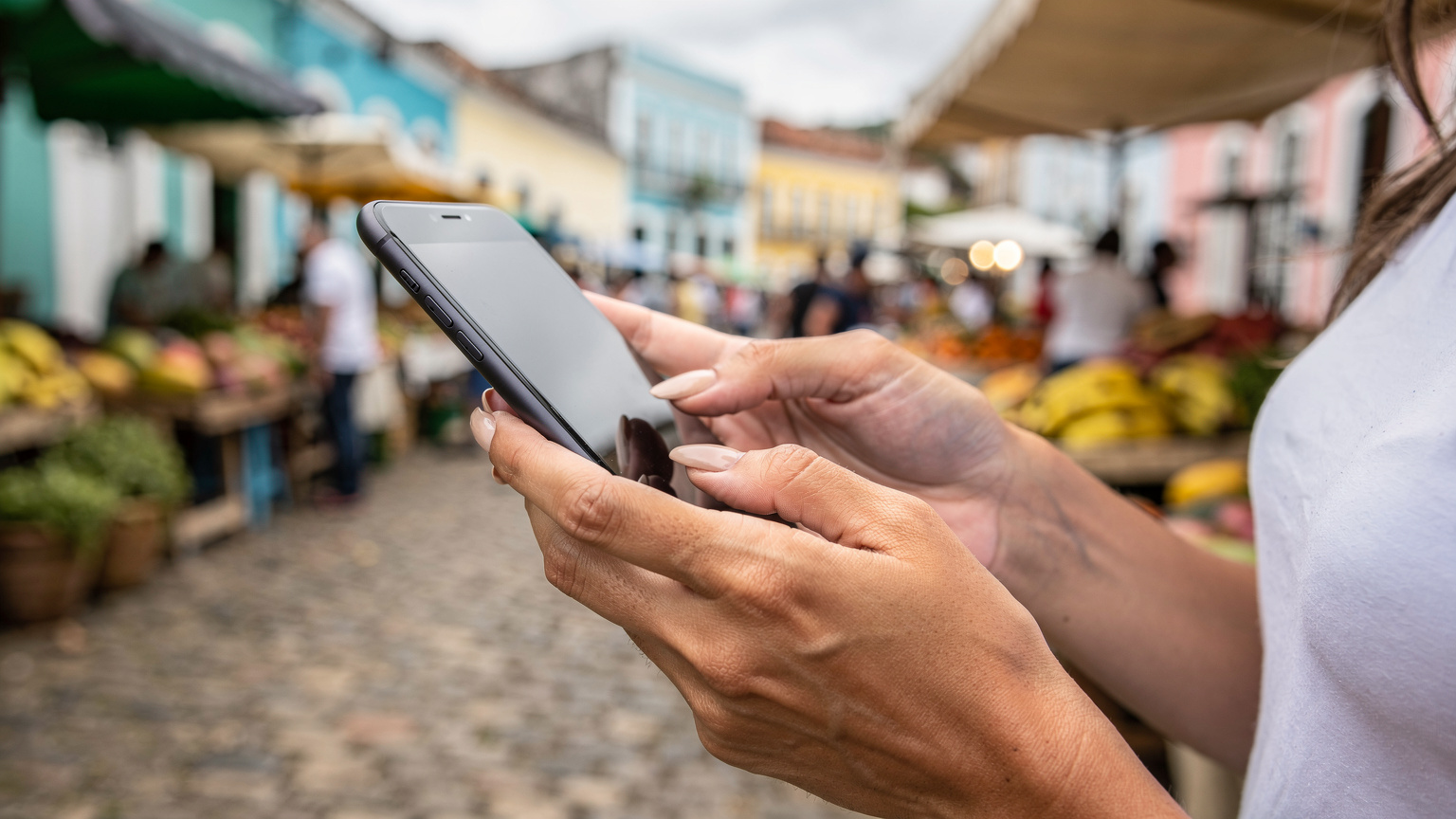 Eine Nahaufnahme zeigt die Hände einer Frau, die vorsichtig ein Smartphone hält und darauf tippt, während sie auf einem Markt in Salvador da Bahia steht.