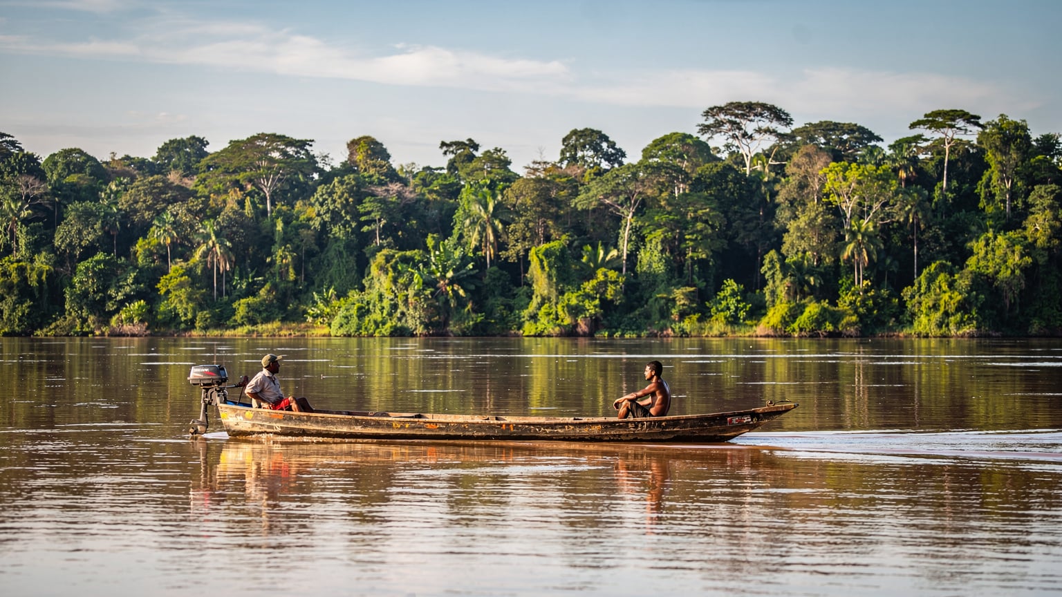 Ein traditionelles Holzboot gleitet auf dem Amazonas in der Nähe von Manaus, umgeben von dichtem Regenwald und einem klaren, weiten Himmel.