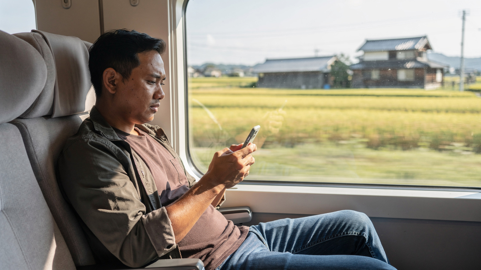 Pemandangan dari dalam kereta Shinkansen yang melaju cepat melewati pedesaan Jepang, dengan seorang penumpang fokus pada ponselnya.