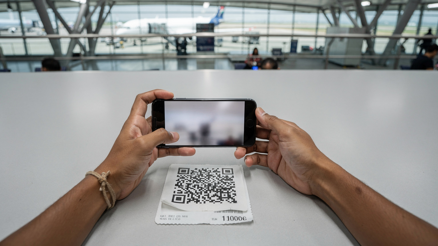 Tangan seorang traveler sedang memindai kode QR eSIM di bandara Incheon, Korea Selatan, dengan latar belakang gerbang keberangkatan.