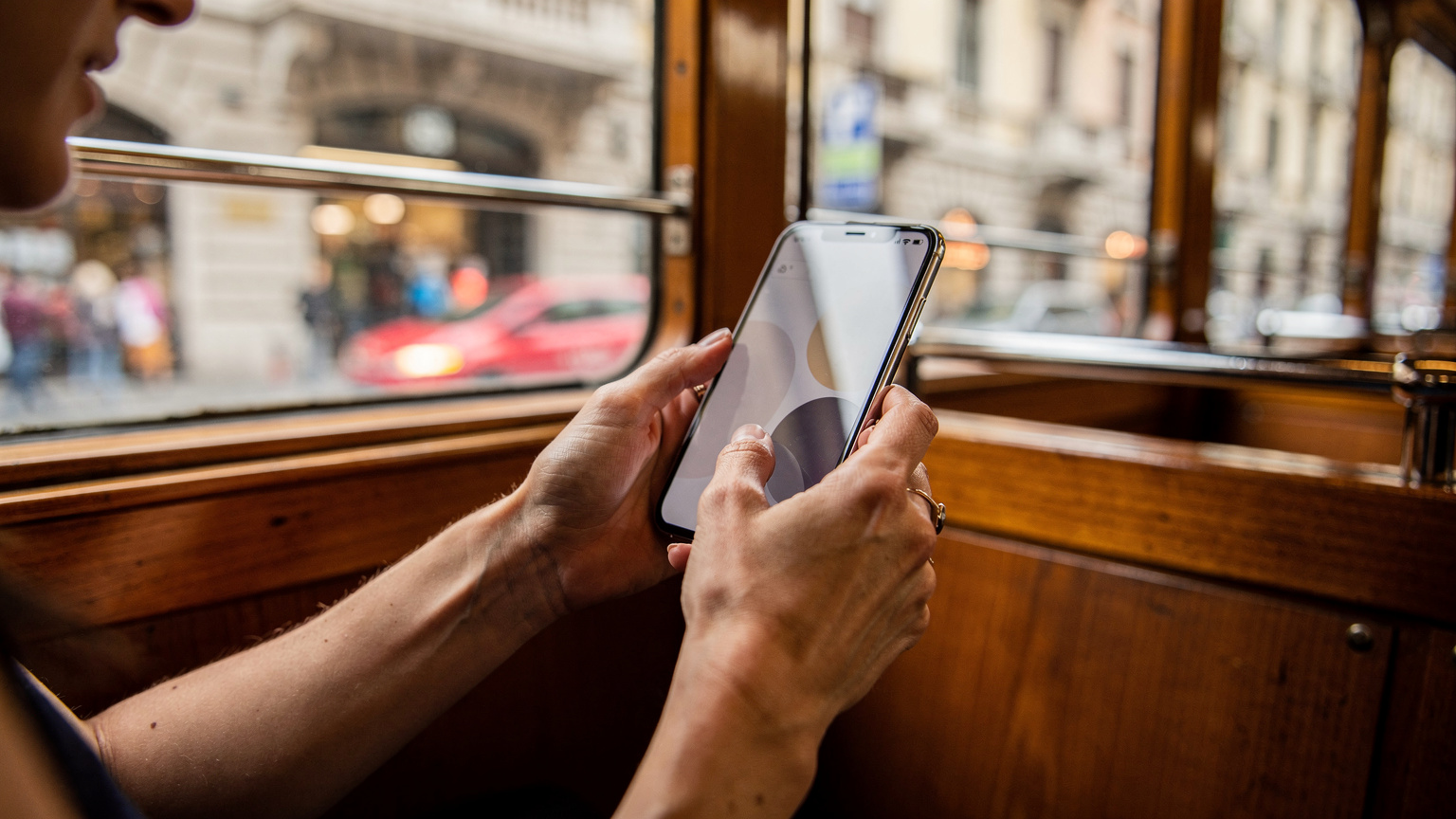Bir kadının elleri, Milano'da tarihi bir tramvayda akıllı telefonunda Cellesim eSIM uygulamasını görüntülüyor. Ekran karartılmış, sadece parmakların hareketi ve uygulamanın silueti belli. Arka planda Milano'nun hareketli sokakları bulanık bir şekilde görülüyor.