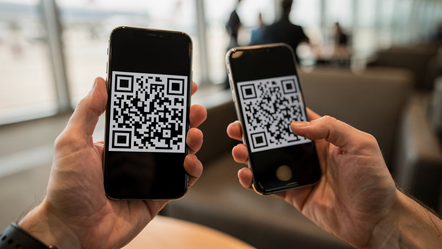 Zbliżenie na dłonie osoby trzymającej smartfon z widocznym kodem QR na ekranie, przygotowującej się do skanowania go innym telefonem, na tle rozmytego wnętrza lotniska.