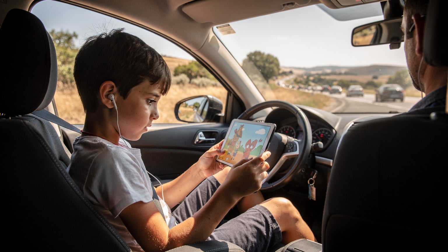 Ein Kind schaut während der Fahrt im Auto, das im Stau steht, auf einem Tablet eine Serie, während die Eltern im Hintergrund leicht gestresst, aber aufmerksam auf die Straße blicken. Die Landschaft draußen deutet auf Südeuropa hin.